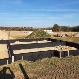 GZB - Kompleksowe prace ziemne, murarskie - Fundamenty w trakcie budowy, zabezpieczone czarną izolacją, z widocznymi stertami piasku i materiałów budowlanych w tle, na tle błękitnego nieba.