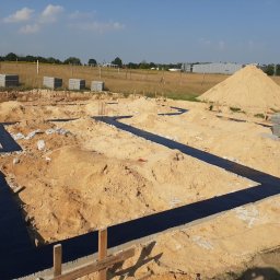 GZB - Kompleksowe prace ziemne, murarskie - Przygotowany teren pod budowę domu z wykopanymi i zabezpieczonymi fundamentami, widoczny stos bloczków betonowych i pryzma piasku na tle łąki i zabudowań gospodarczych.