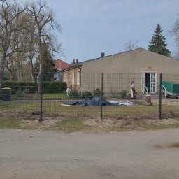 GZB - Kompleksowe prace ziemne, murarskie - Montaż ogrodzenia panelowego wokół posesji z budynkiem, widoczne słupki i przęsła, przygotowany teren z wykopaną ziemią, w tle budynek z przyczepą pod plandeką i pracownik w jasnych ubraniach.