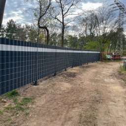 GZB - Kompleksowe prace ziemne, murarskie - Nowoczesne ogrodzenie panelowe z poziomym, białym pasem, otoczone zielenią i ziemistą ścieżką, widok z perspektywy gruntu, słoneczny dzień.