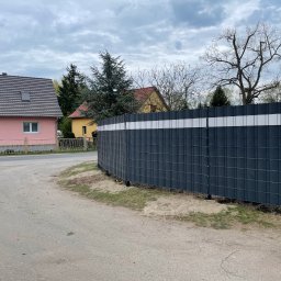 GZB - Kompleksowe prace ziemne, murarskie - Nowoczesne ogrodzenie panelowe w kolorze grafitowym z białym pasem, otoczone zielenią i domami w tle, prezentuje estetyczne oddzielenie przestrzeni.