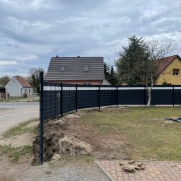 GZB - Kompleksowe prace ziemne, murarskie - Nowoczesne, czarne ogrodzenie panelowe z białymi akcentami, oddzielające posesję. Widoczny fragment trawnika, domy w tle i pochmurne niebo.