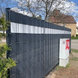 GZB - Kompleksowe prace ziemne, murarskie - Nowoczesne ogrodzenie panelowe z wypełnieniem z tworzywa sztucznego w kolorze grafitowym, z białym pasem na górze. Na tle budynek mieszkalny i drzewa.