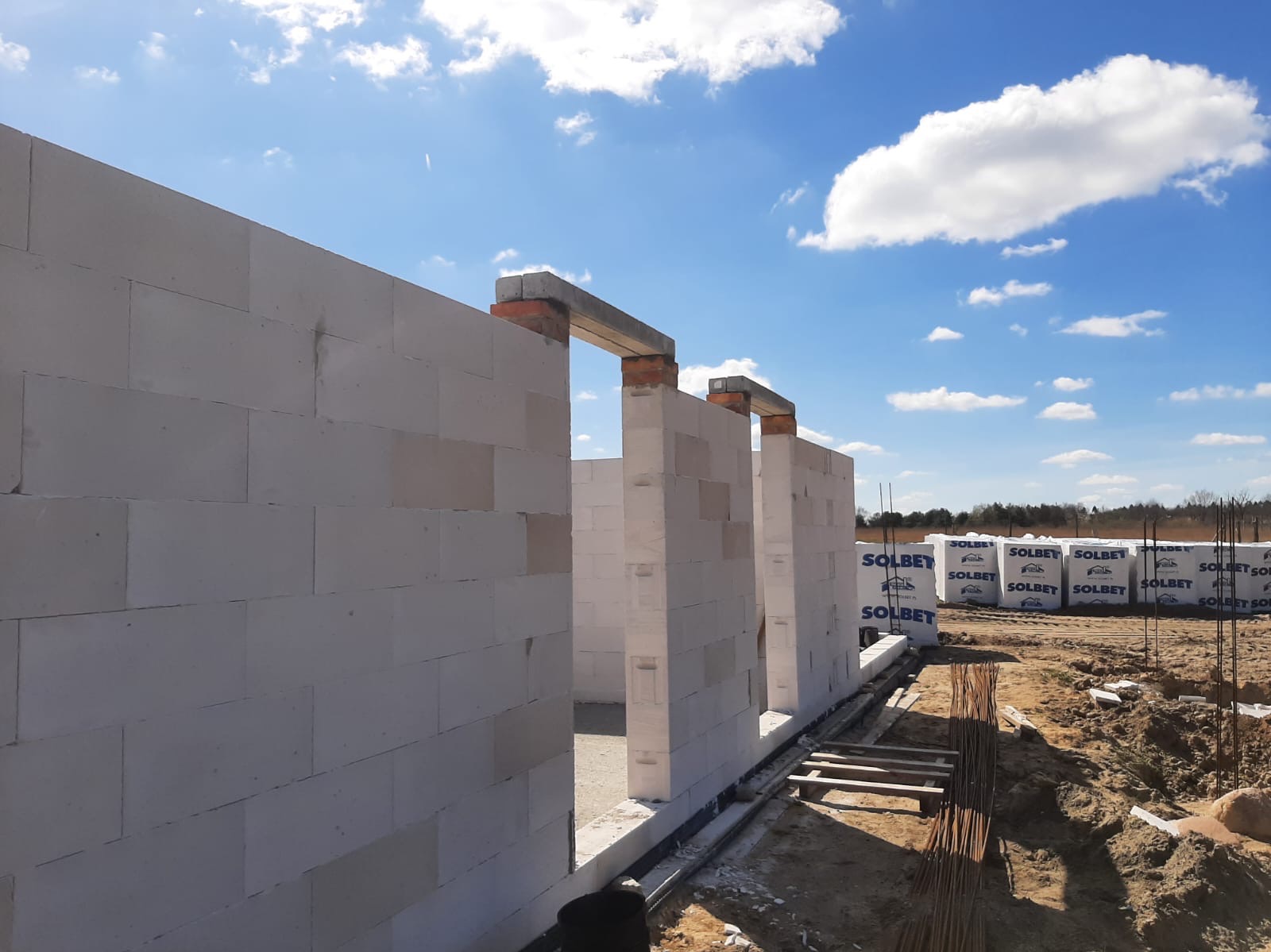 Wznoszenie ścian z białych bloczków betonu komórkowego Solbet na fundamencie, widoczne zbrojenie i palety z materiałem budowlanym w tle, błękitne niebo z chmurami.