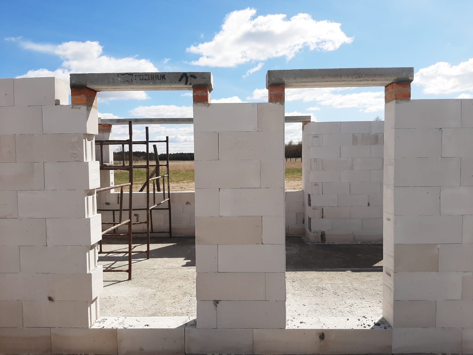 Wznoszone ściany domu z betonu komórkowego Ytong, widoczne nadproża z napisem 'POZBRUK' i metalowe rusztowanie wewnątrz, w tle łąka pod błękitnym niebem.