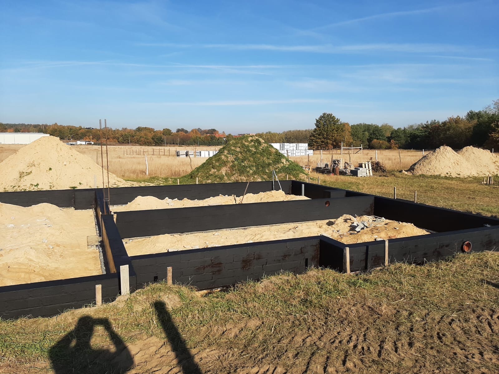 Fundamenty w trakcie budowy, zabezpieczone czarną izolacją, z widocznymi stertami piasku i materiałów budowlanych w tle, na tle błękitnego nieba.