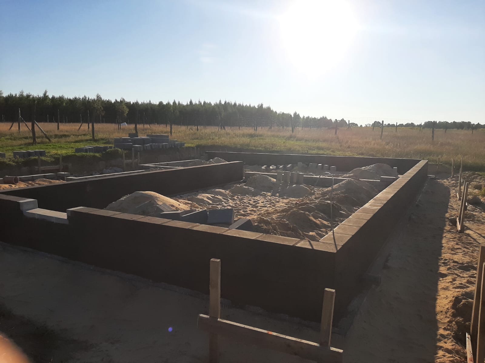 Fundamenty pod budynek z bloczków betonowych, wypełnione piaskiem, na tle łąki i lasu w słoneczny dzień.