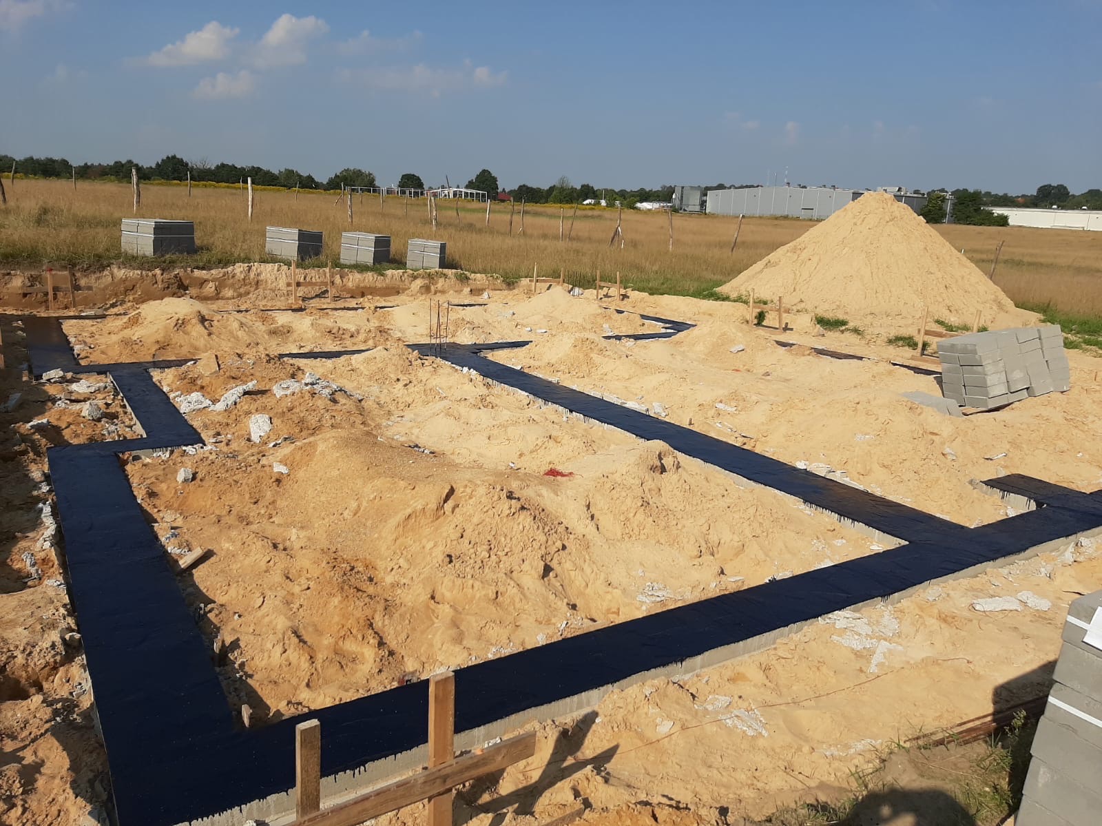 Przygotowany teren pod budowę domu z wykopanymi i zabezpieczonymi fundamentami, widoczny stos bloczków betonowych i pryzma piasku na tle łąki i zabudowań gospodarczych.