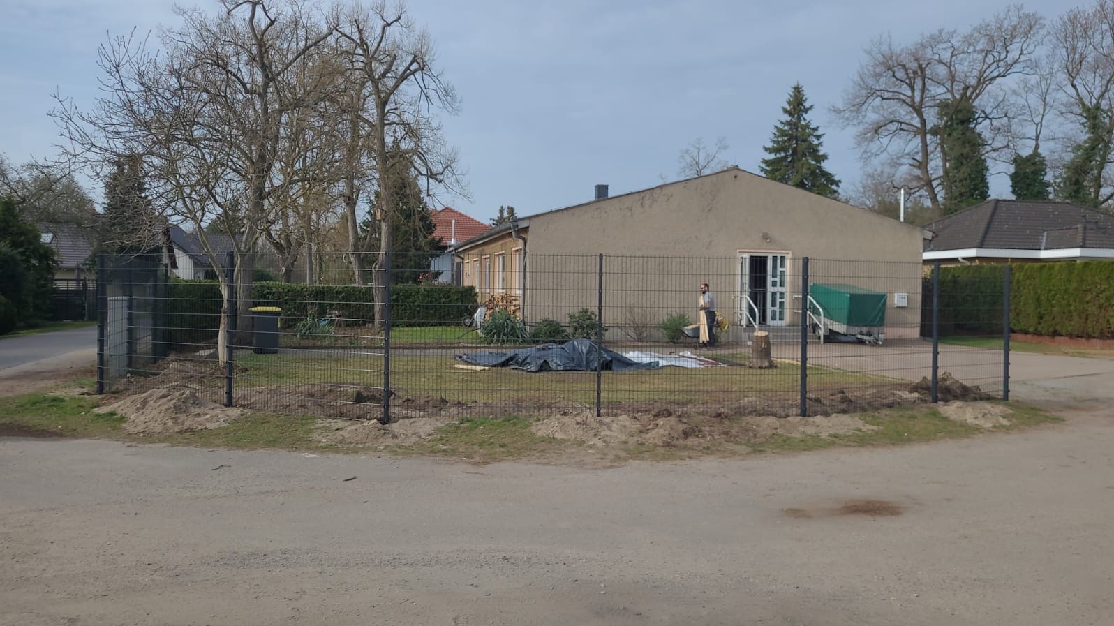 Montaż ogrodzenia panelowego wokół posesji z budynkiem, widoczne słupki i przęsła, przygotowany teren z wykopaną ziemią, w tle budynek z przyczepą pod plandeką i pracownik w jasnych ubraniach.