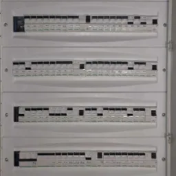 Rozdzielnia elektryczna z wieloma bezpiecznikami, widok z bliska, biała obudowa, czarne przełączniki, oznaczenia na bezpiecznikach, przewody w górnej części.