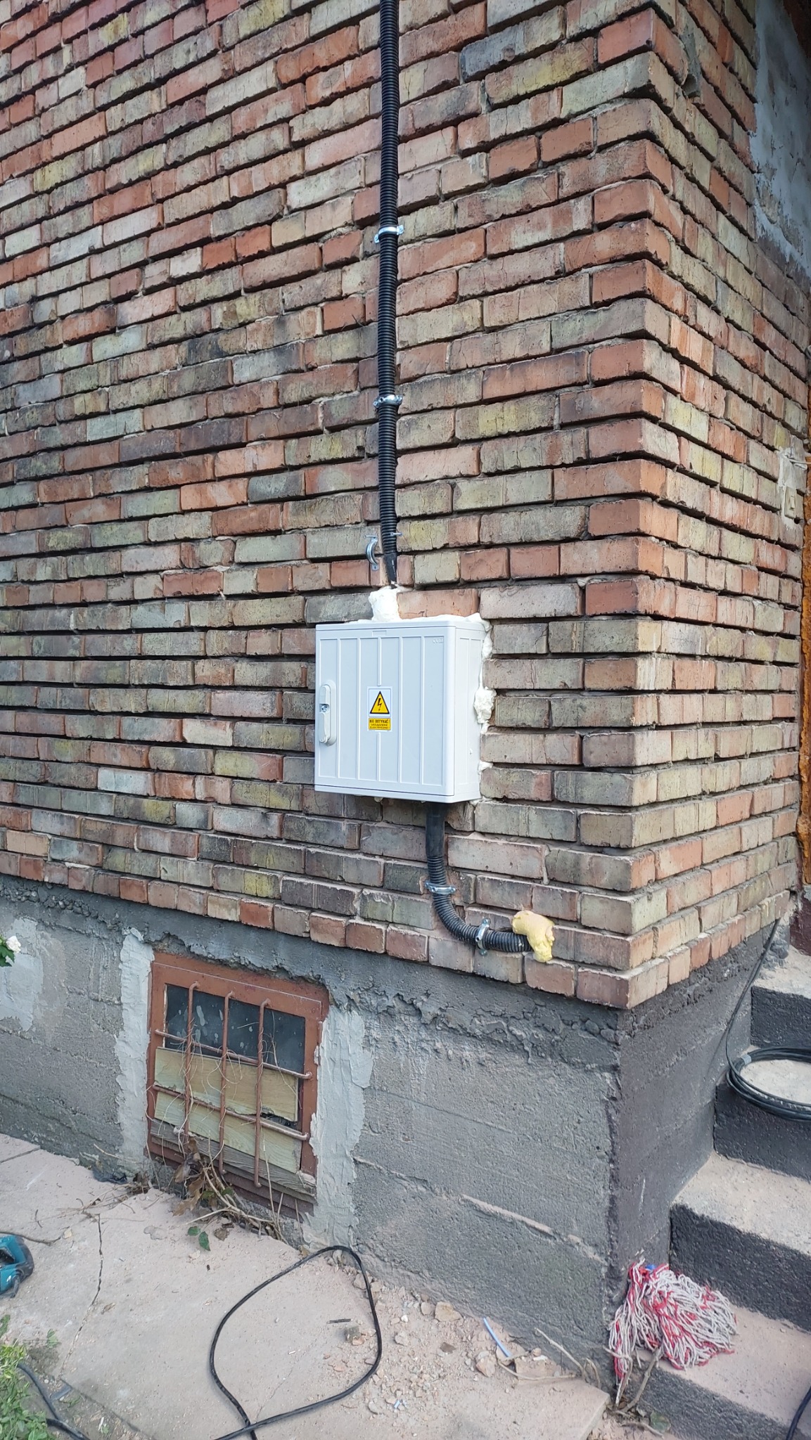 Zewnętrzna skrzynka elektryczna z żółtą naklejką ostrzegawczą o wysokim napięciu, zamontowana na ceglanej ścianie budynku, z widocznymi rurami osłonowymi i śladami pianki montażowej.