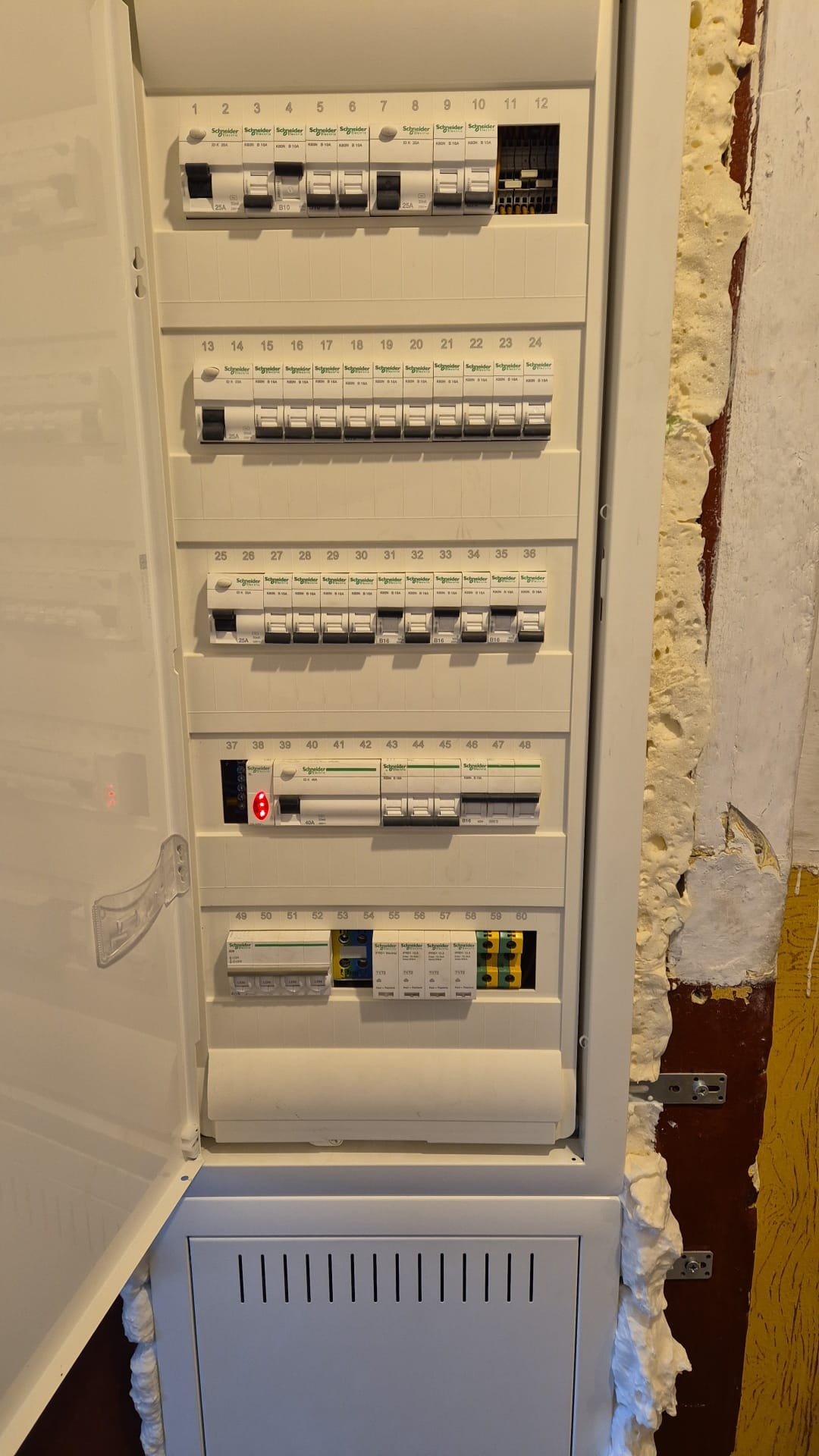 Otwarta skrzynka z bezpiecznikami, widoczne wyłączniki automatyczne marki Schneider, instalacja w trakcie montażu z widoczną pianką montażową wokół obudowy.