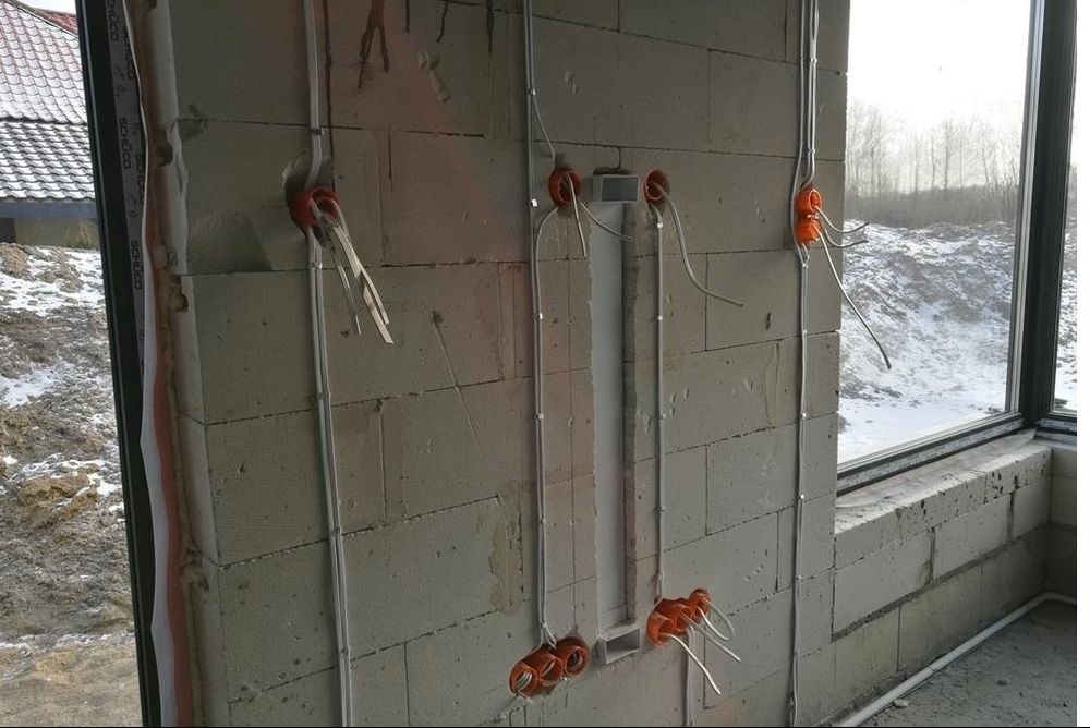 Surowy mur z betonu komórkowego z widocznymi puszkami instalacyjnymi i przewodami elektrycznymi w trakcie montażu, widok przez okno na ośnieżony teren.