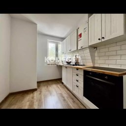 Mazur.s.m - Jasna kuchnia z białymi szafkami, drewnianym blatem i panelami podłogowymi. Widoczna czarna płyta grzewcza i piekarnik. Ściany pomalowane na biało, a nad blatem białe, prostokątne płytki. W tle...