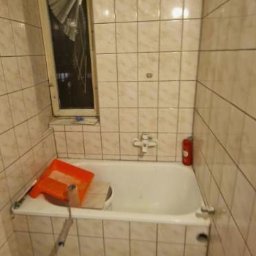 Mazur.s.m - Łazienka w trakcie remontu, widoczna wanna z akcesoriami malarskimi, stare płytki na ścianach i podłodze, uszkodzona żaluzja w oknie, ślady po naprawach na suficie.