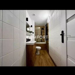 Mazur.s.m - Wąska łazienka z białymi kwadratowymi płytkami, drewnianą podłogą i szafką pod umywalką; widoczny fragment kabiny prysznicowej za toaletą.