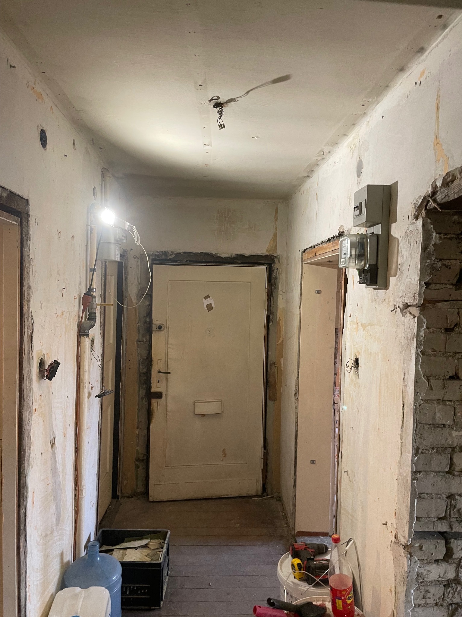 Korytarz w trakcie remontu: ściany z resztkami starej farby i tapet, odsłonięte przewody elektryczne, prowizoryczne oświetlenie halogenowe, widoczne narzędzia i materiały budowlane, stara stolarka...