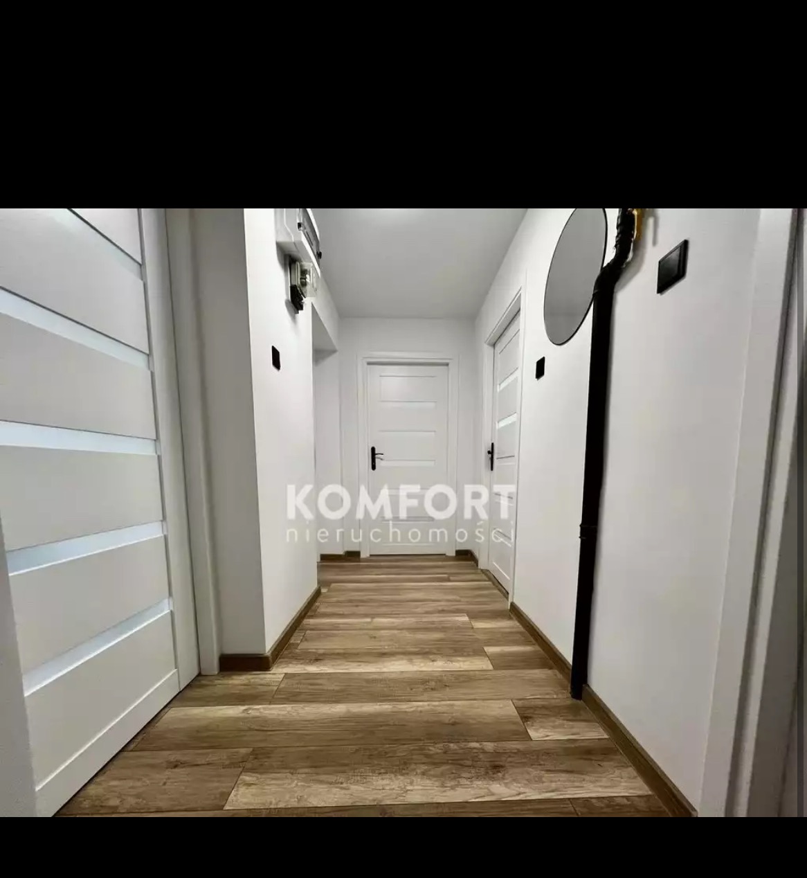Wnętrze korytarza z panelami podłogowymi imitującymi drewno, białymi ścianami i drzwiami, widoczne elementy instalacji elektrycznej i wodnej, logo firmy 'Komfort Nieruchomości'.
