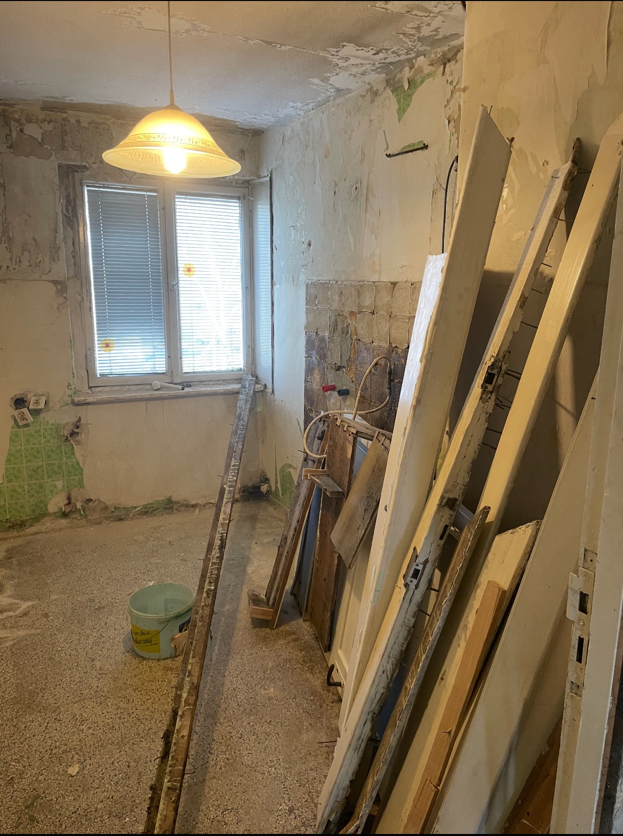 Pokój w trakcie remontu: zerwana tapeta, odsłonięte fragmenty starej glazury, przygotowane do malowania drewniane elementy, wiadro z farbą na podłodze.