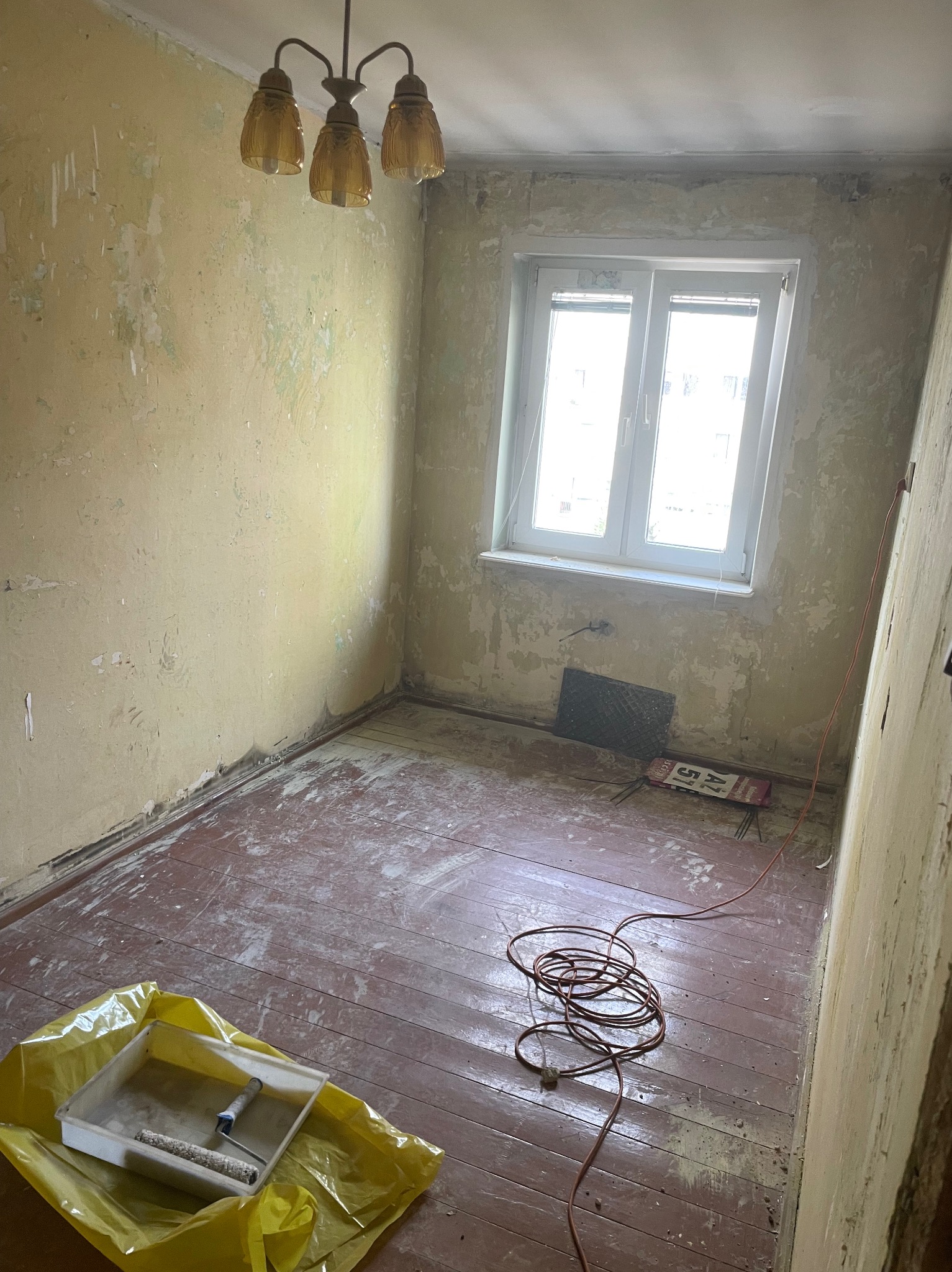 Pokój przed malowaniem: zniszczone ściany z resztkami farby, drewniana podłoga z plamami, okno z widokiem na zewnątrz, żółta folia malarska z kuwetą i wałkiem, zwinięty kabel na podłodze, lampa...