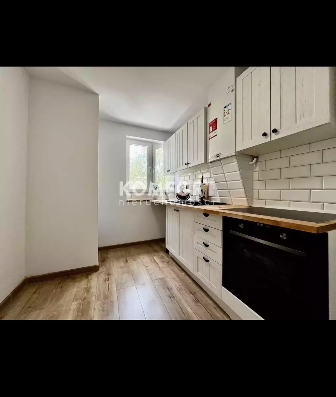 Jasna kuchnia z białymi szafkami, drewnianym blatem i panelami podłogowymi. Widoczna czarna płyta grzewcza i piekarnik. Ściany pomalowane na biało, a nad blatem białe, prostokątne płytki. W tle...