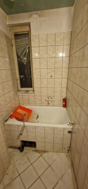 Łazienka w trakcie remontu, widoczna wanna z akcesoriami malarskimi, stare płytki na ścianach i podłodze, uszkodzona żaluzja w oknie, ślady po naprawach na suficie.