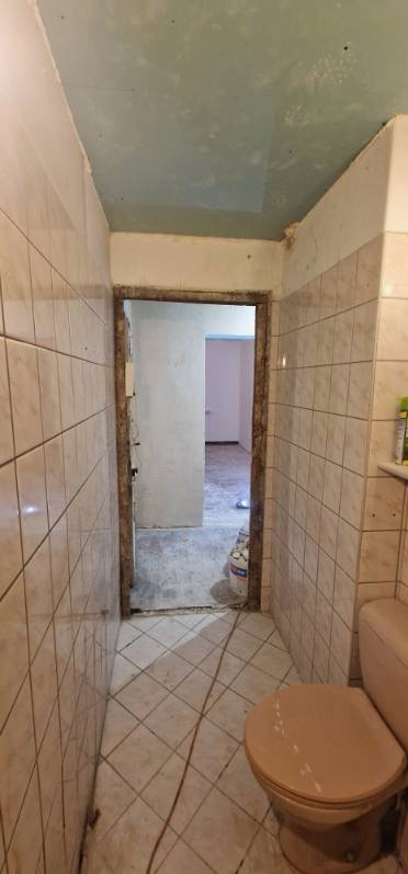 Łazienka w trakcie remontu: stare, beżowe płytki na ścianach i podłodze, widoczny surowy stan ściany nad drzwiami, niedomalowany sufit i toaleta w stylu retro.