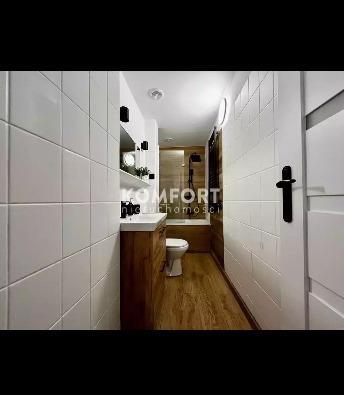 Wąska łazienka z białymi kwadratowymi płytkami, drewnianą podłogą i szafką pod umywalką; widoczny fragment kabiny prysznicowej za toaletą.