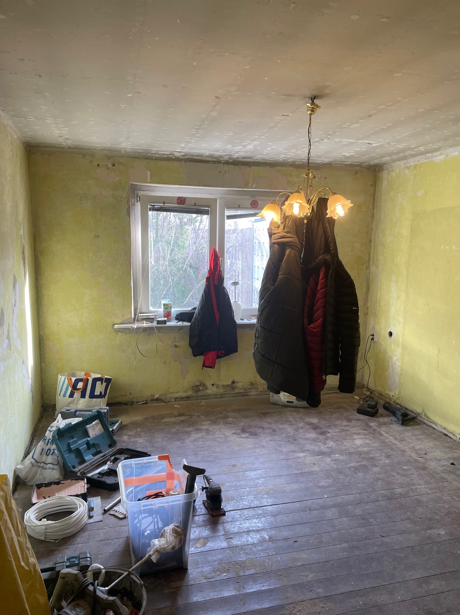 Pokój w trakcie remontu: ściany z resztkami farby, narzędzia malarskie, wiszące kurtki, stara drewniana podłoga.