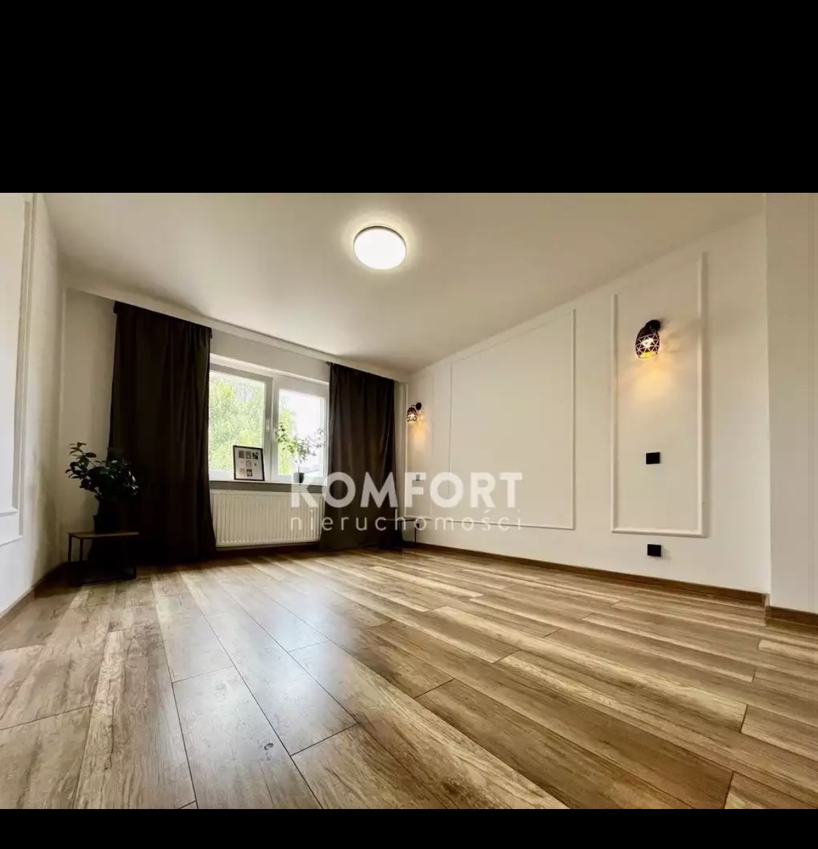 Jasne, puste pomieszczenie z panelami podłogowymi w odcieniach brązu, białymi ścianami z dekoracyjnymi listwami, brązowymi zasłonami i dwoma kinkietami z czarnymi elementami. Widoczne logo firmy...