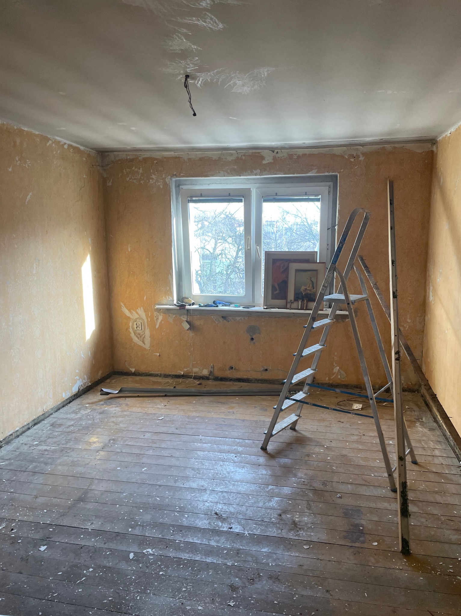 Pokój przed remontem: widoczne ślady po starych tapetach na ścianach, drewniana podłoga z resztkami farby, drabina oparta o ścianę, okno z widokiem na drzewa, obrazy oparte o parapet.