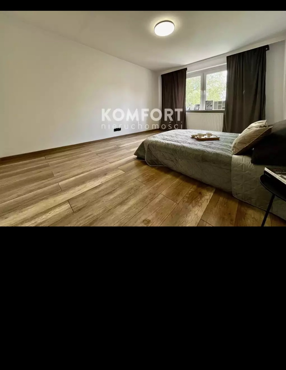 Jasne wnętrze sypialni z drewnopodobną podłogą, szarym łóżkiem i ciemnymi zasłonami, widoczne logo 'Komfort nieruchomości' na ścianie.