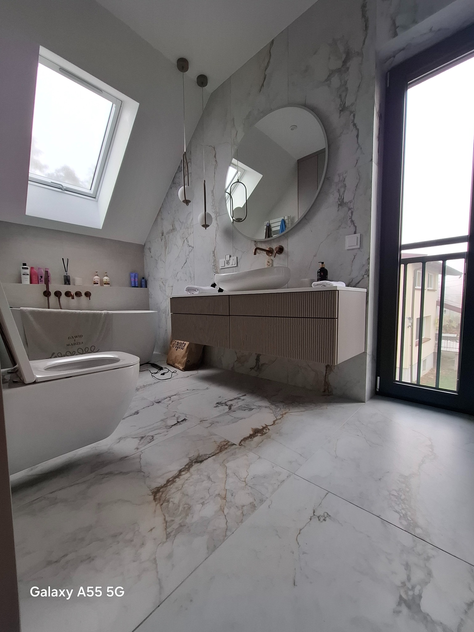 Nowoczesna łazienka na poddaszu z marmurowymi płytkami, owalnym lustrem i wiszącą szafką z umywalką, widok na okno dachowe i balkon.