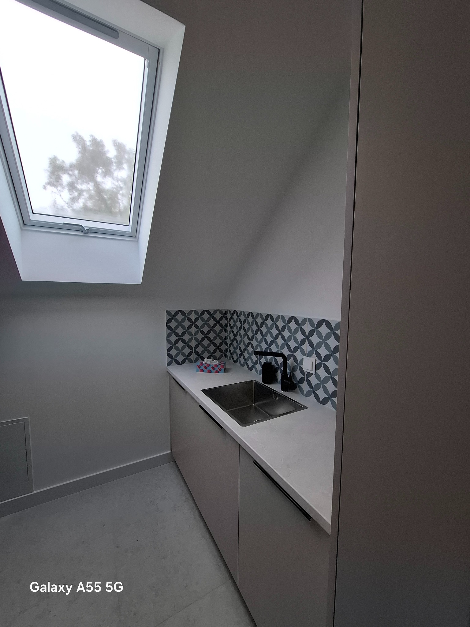 Umywalka w szafce z czarną armaturą i dekoracyjnymi płytkami na ścianie w łazience na poddaszu z oknem dachowym.