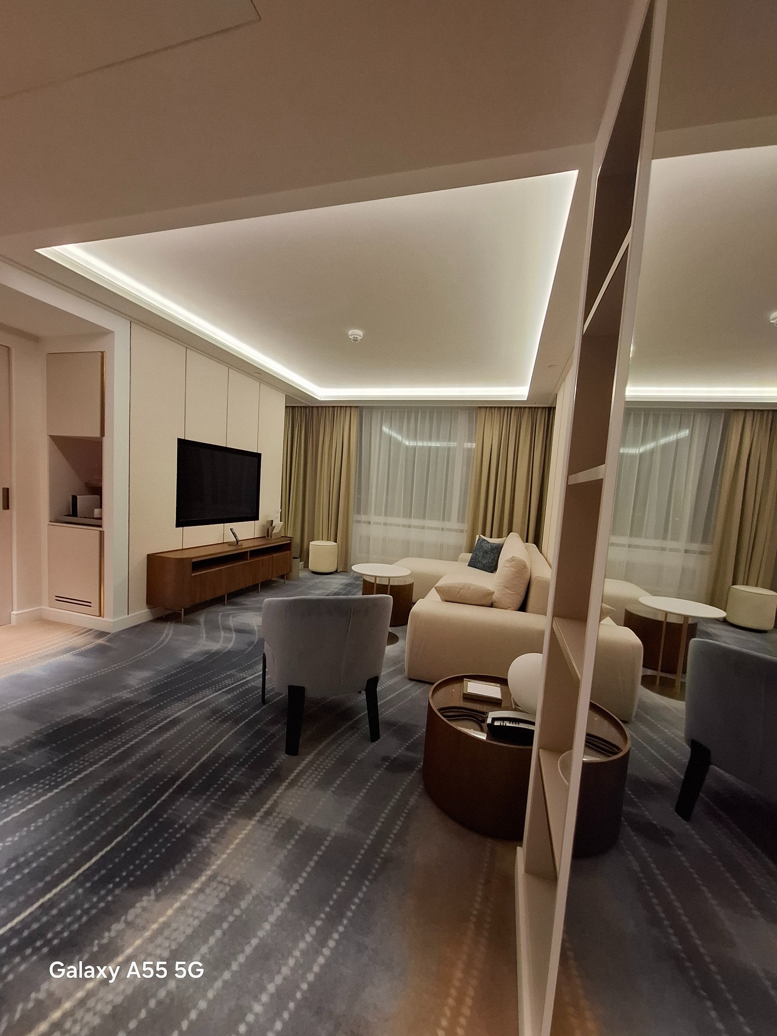 Nowoczesny salon z oświetleniem LED w suficie, beżową sofą z poduszkami, szarym dywanem w paski i telewizorem na drewnianej szafce, widziany z perspektywy niskiego kąta.