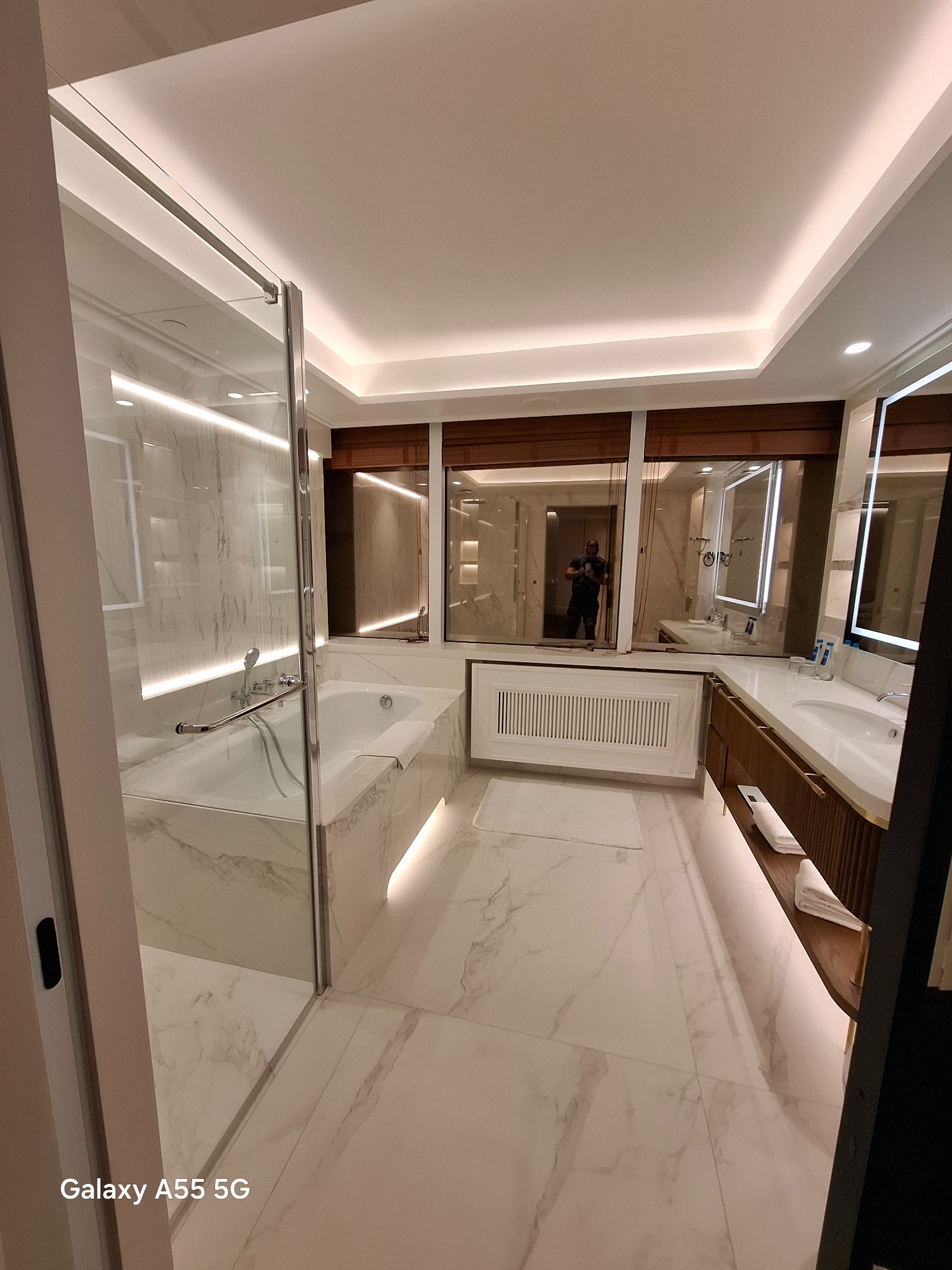 Elegancka łazienka z marmurowymi płytkami, oświetleniem LED pod wanną i szafkami, przeszkloną kabiną prysznicową i dużym lustrem z podświetleniem, w odbiciu widać osobę robiącą zdjęcie.