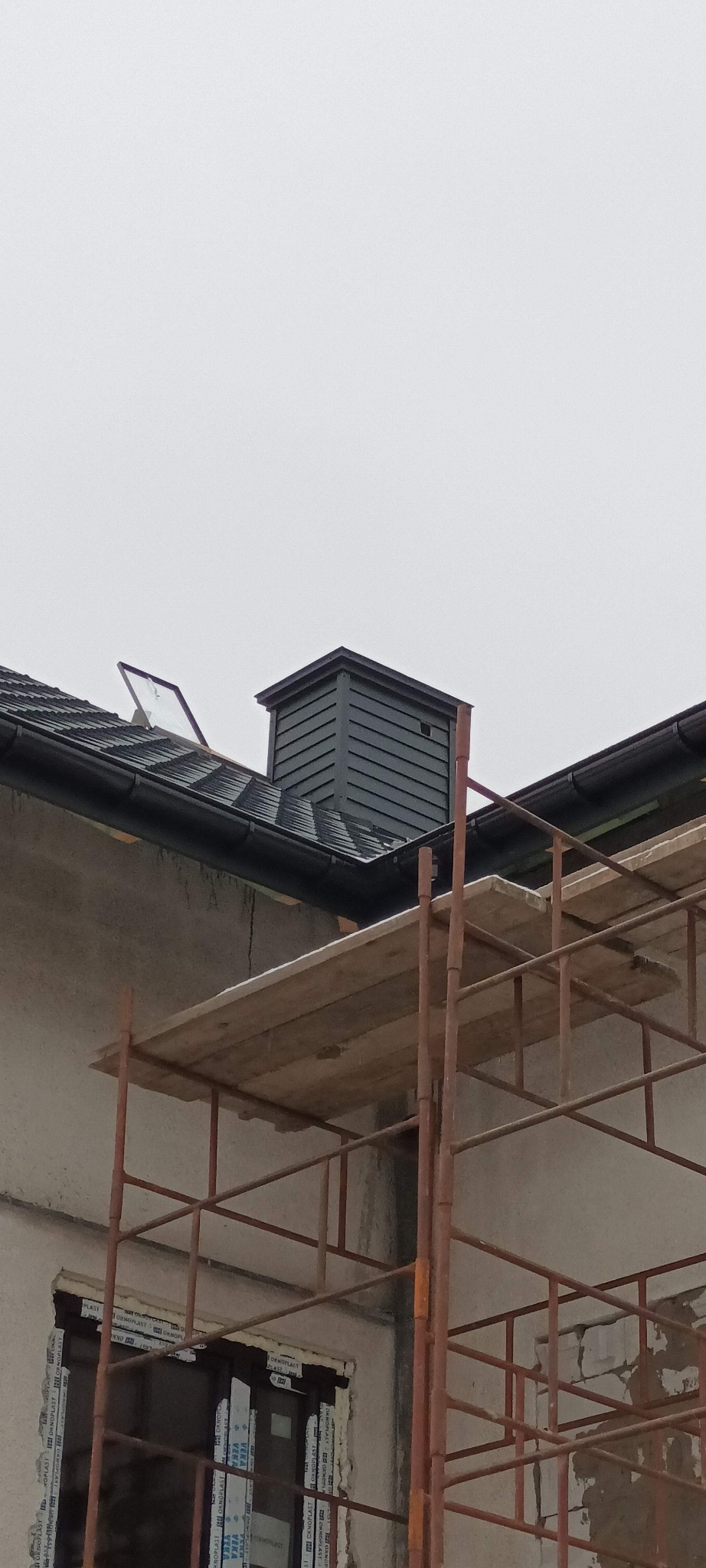 Szary dach z oknem dachowym i kominem w trakcie budowy, widoczne rusztowanie. Ujęcie z dołu, pochmurne niebo w tle. Detal z budowy dachu.