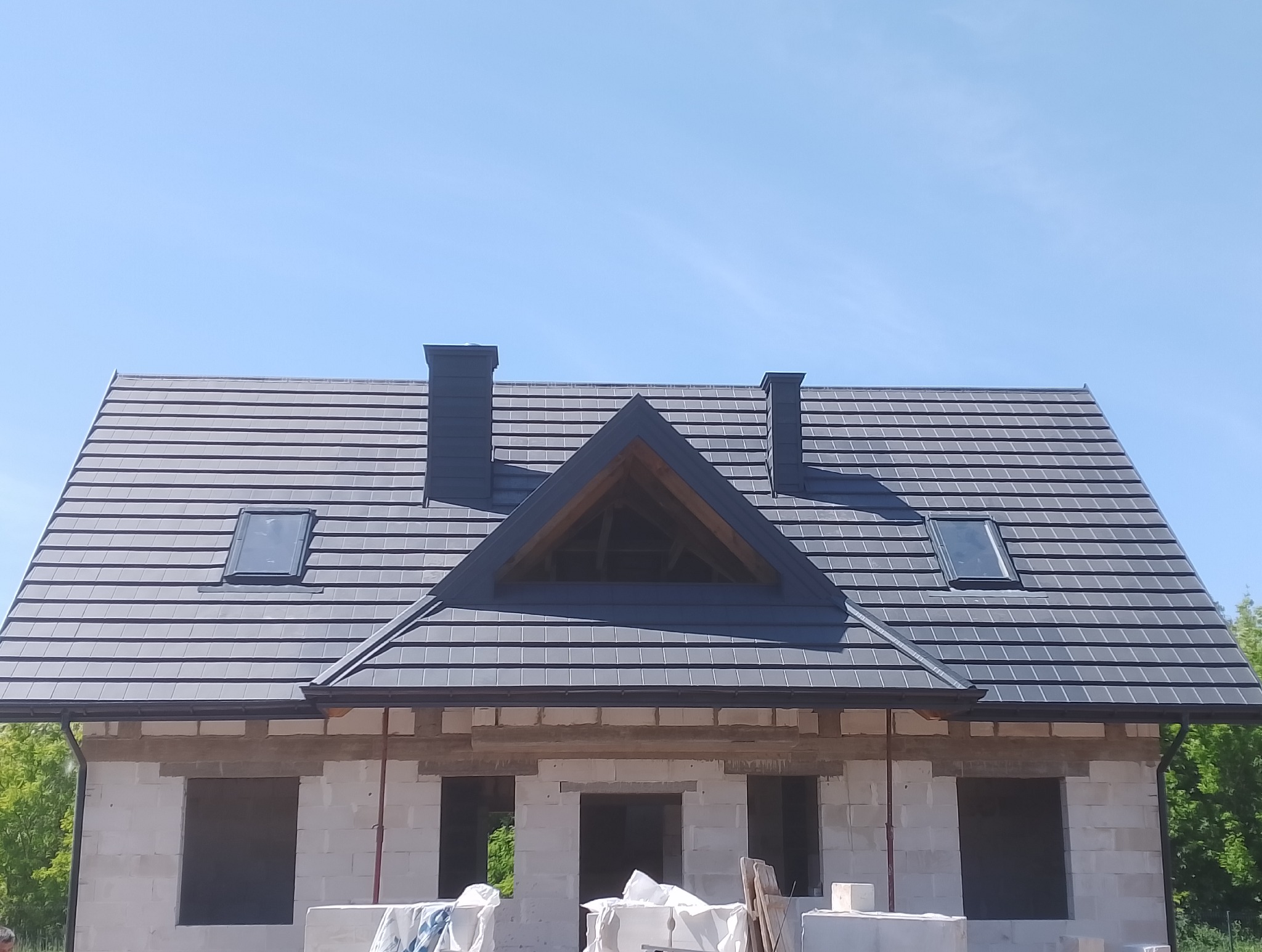 Nowoczesny dom w trakcie budowy z ciemnoszarą dachówką, oknami dachowymi i dwoma kominami. Widoczna surowa konstrukcja ścian z białego bloczka.