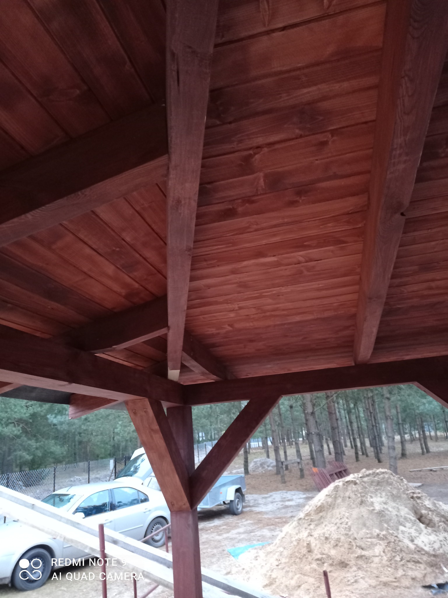 Drewniana konstrukcja z belek i desek, prawdopodobnie zadaszenie lub altana w trakcie budowy. Widoczne elementy konstrukcyjne i otoczenie budowy.