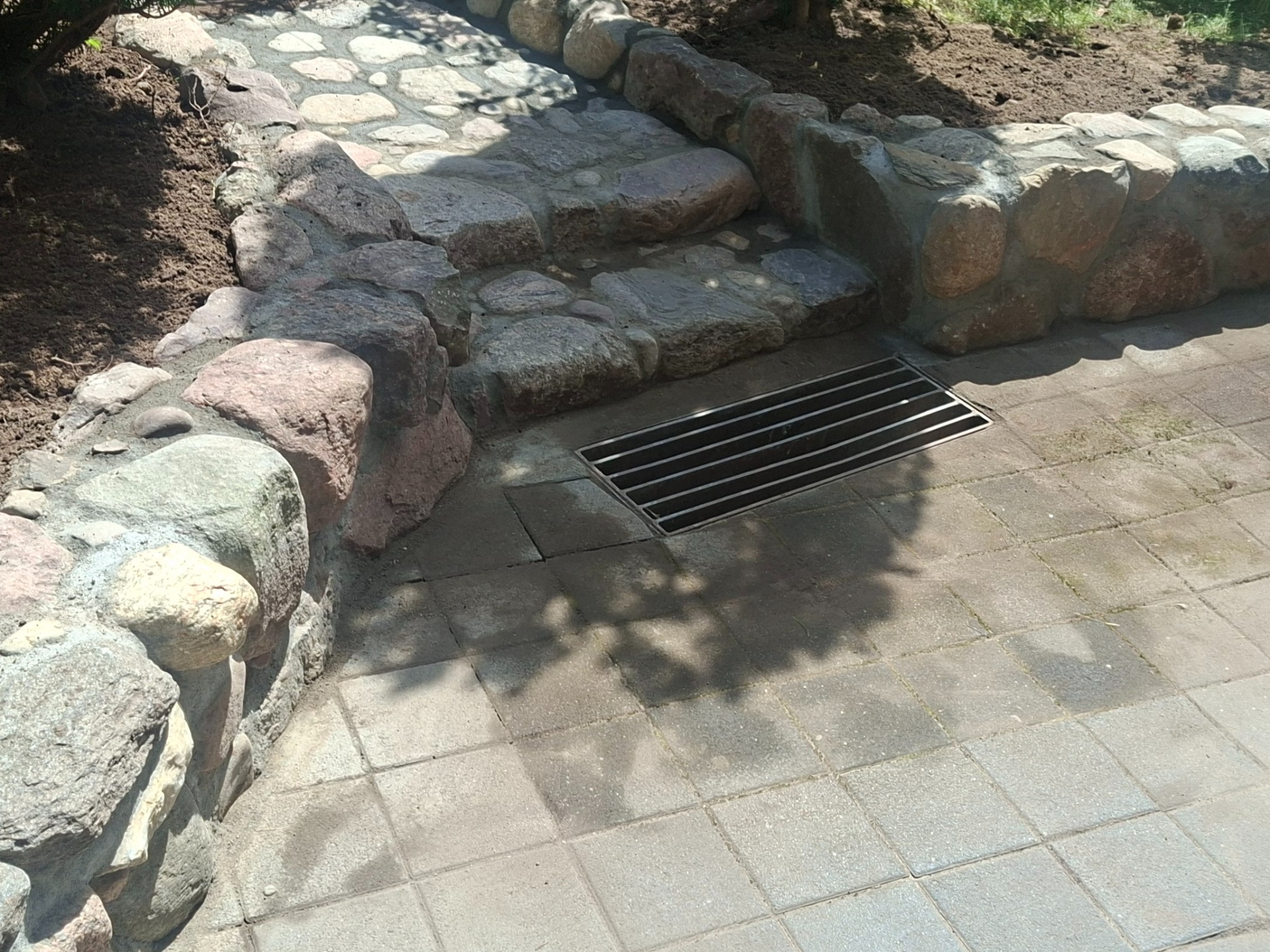 Kamienny murek z polnego kamienia, schody i chodnik z betonowej kostki. Na chodniku widoczna studzienka kanalizacyjna z metalową kratką.