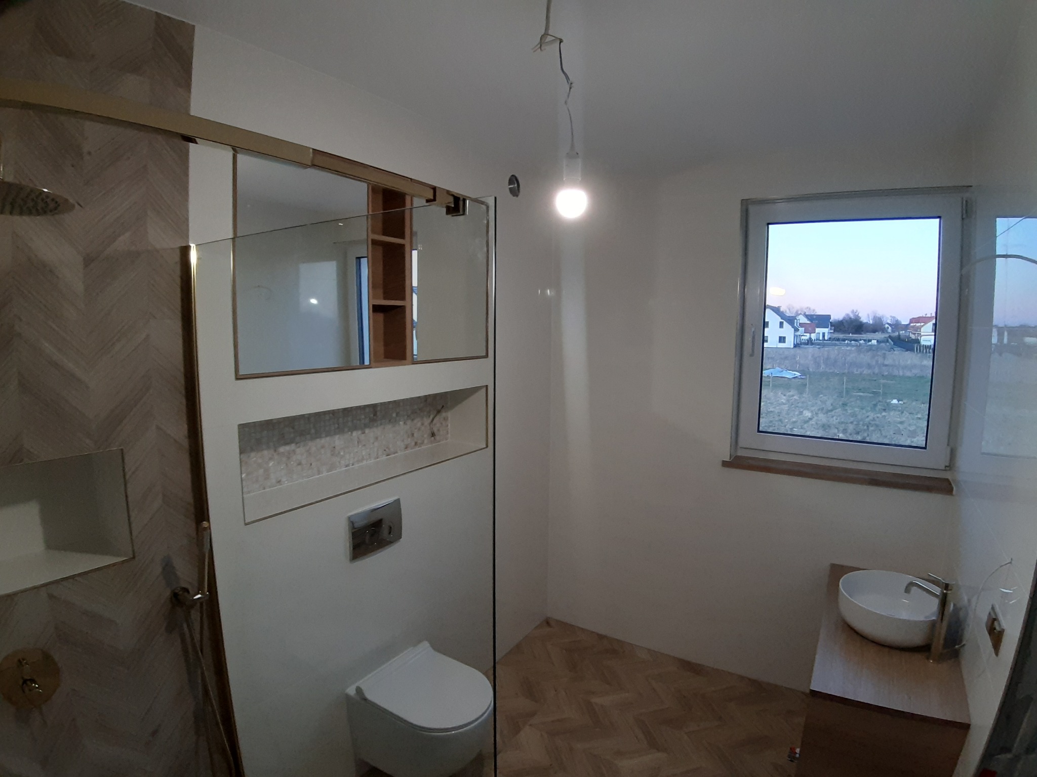 Nowoczesna łazienka po remoncie z kabiną prysznicową typu walk-in, umywalką nablatową i oknem z widokiem na zewnątrz. Uwagę zwraca wzór parkietu na podłodze.