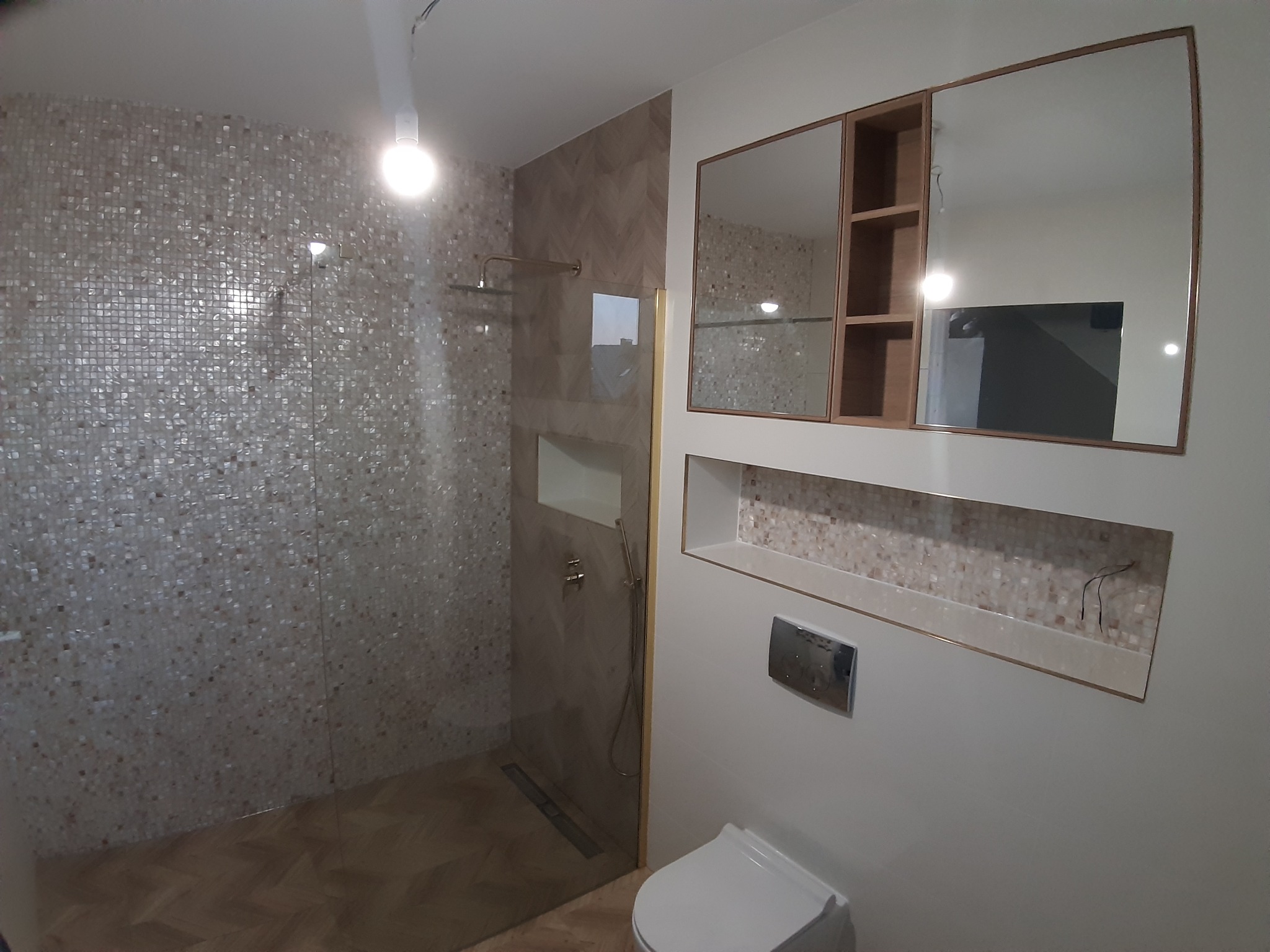 Nowoczesna łazienka po remoncie: kabina prysznicowa z mozaiką, toaleta, szafka z lustrem. Wykończenie w beżach i bieli, z akcentami złota. Widoczne detale wykończeniowe.