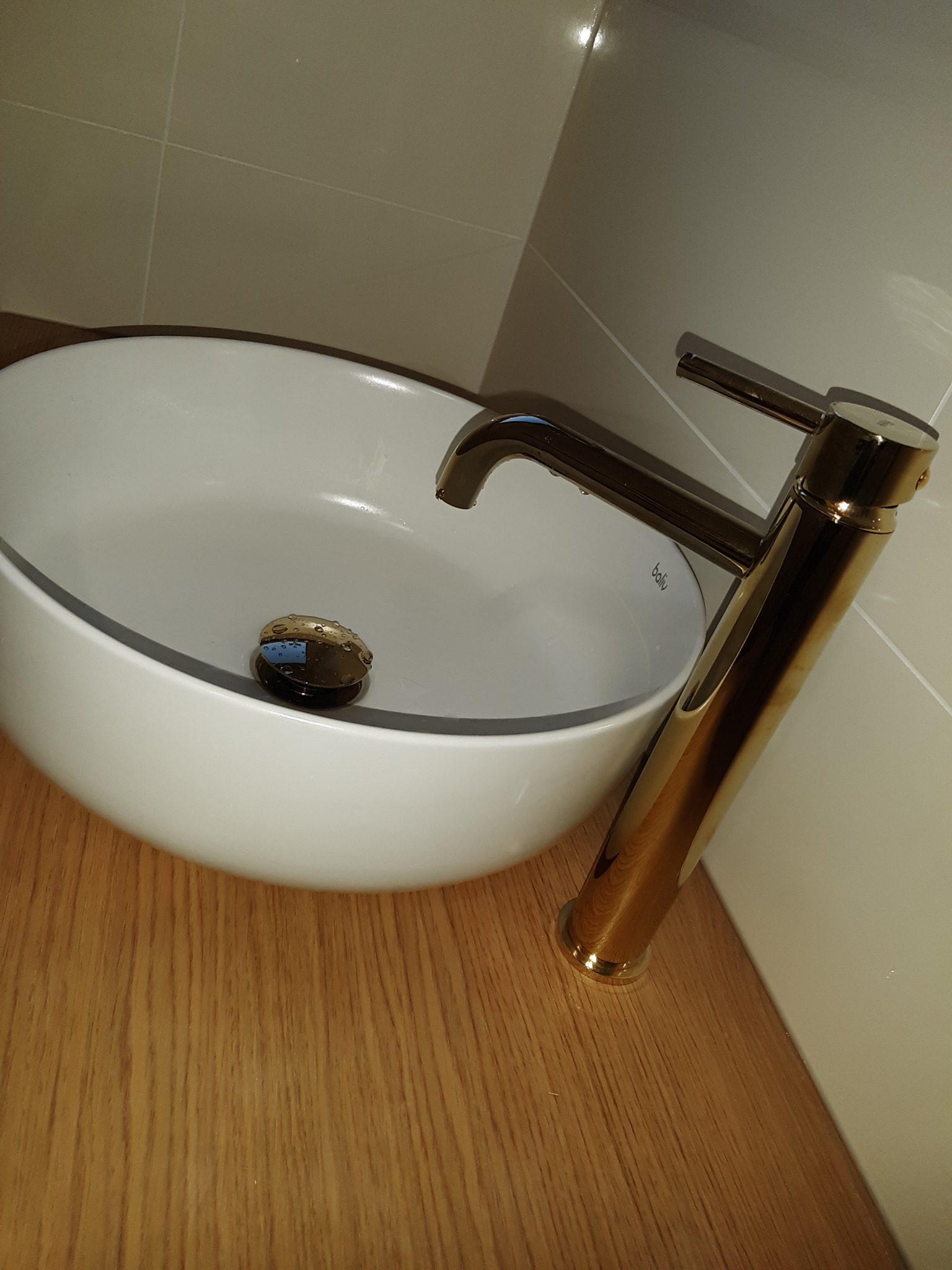 Biała umywalka nablatowa z złotą baterią o nowoczesnym designie, zamontowana na drewnianym blacie w łazience z beżowymi płytkami.