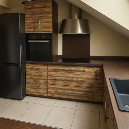 Luca Design - Zabudowa kuchenna z forniru w odcieniach brązu, z czarną lodówką i okapem w nowoczesnym wnętrzu ze skośnym sufitem. Widoczny fragment zlewu.