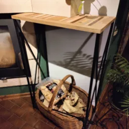 Konsola z blatem z mozaiki drewna na metalowych nogach, pod nią kosz z drewnem kominkowym. W tle kominek i rośliny doniczkowe.