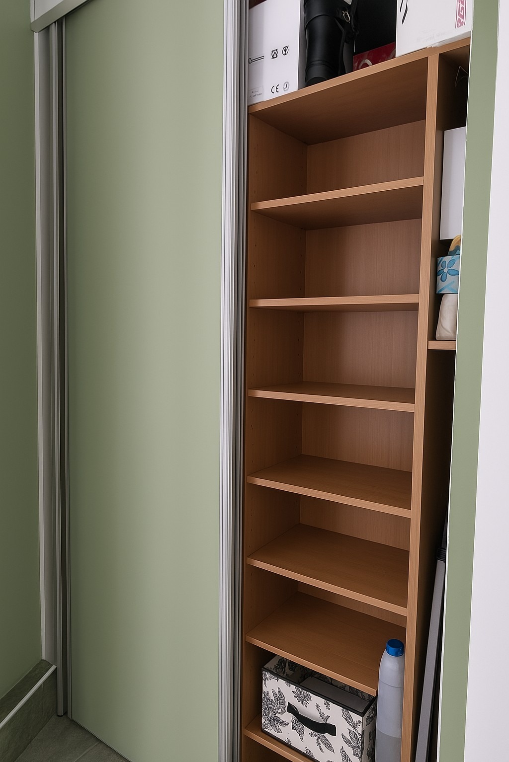 Otwarta szafa wnękowa z przesuwnymi drzwiami w odcieniach zieleni. Półki z jasnego drewna, na nich pudełka i przedmioty. Minimalistyczny design, stonowana kolorystyka.