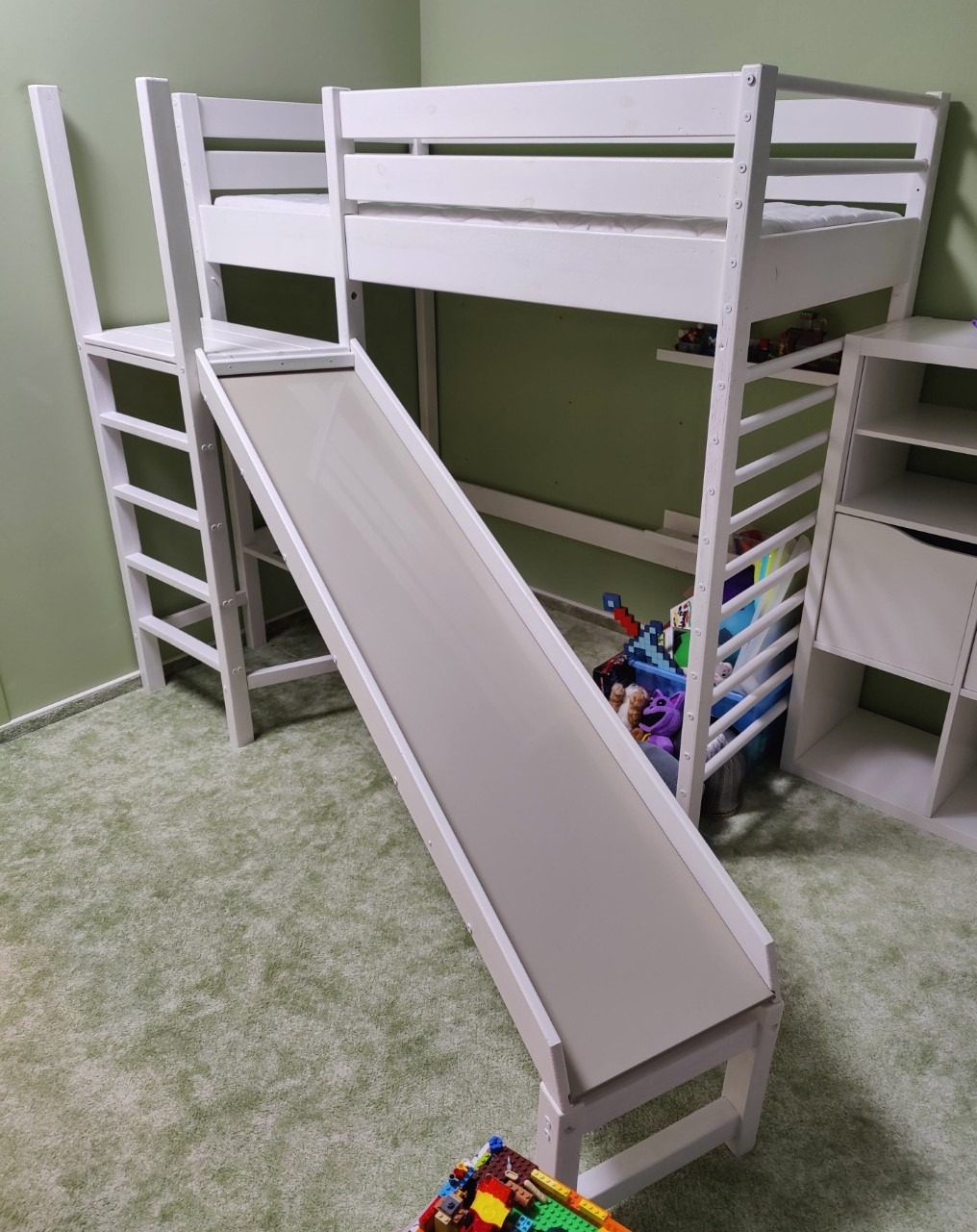 Białe łóżko piętrowe z drabinką i zjeżdżalnią w pokoju dziecięcym z zieloną wykładziną. Pod łóżkiem widoczne zabawki i biały regał.