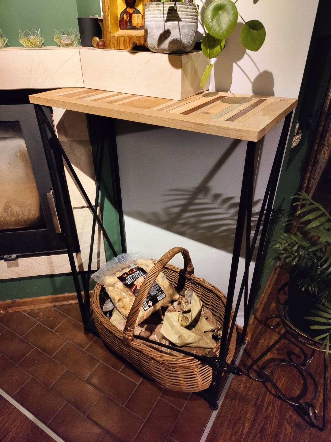 Konsola z blatem z mozaiki drewna na metalowych nogach, pod nią kosz z drewnem kominkowym. W tle kominek i rośliny doniczkowe.