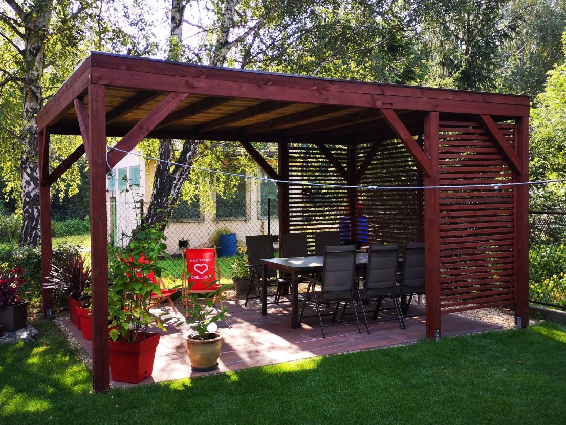 Drewniana pergola z zadaszeniem i ażurową ścianą boczną, wyposażona w stół i krzesła. Czerwone donice z roślinami i krzesło ogrodowe dodają koloru.