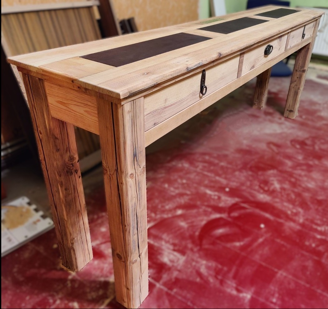 Długi, smukły stół z jasnego drewna z trzema szufladami, każda z ciemnym, prostokątnym blatem. Widoczne słoje drewna i metalowe uchwyty szuflad.